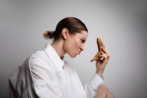 Portrait of young angry woman wearing white shirt and holding a ripe yellow banana, making scared grimace. - 写真・画像