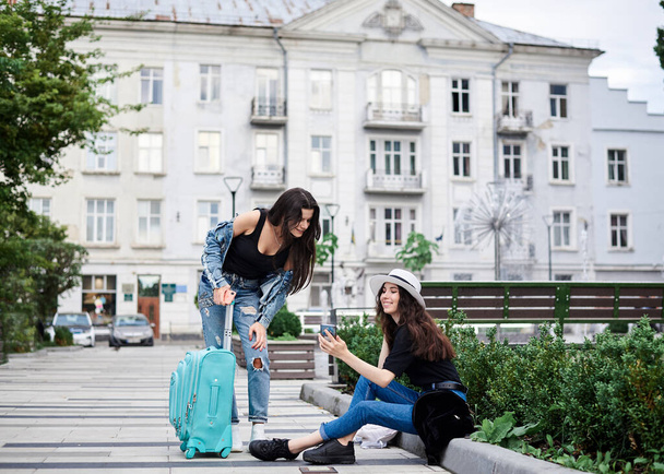 Two young brunette girls, wearing casual jeans clothes, with mint luggage, sitting on pavement border, checking phone in front of historical building in town. City sightseeing tour.Tourism lifestyle. - Foto, afbeelding