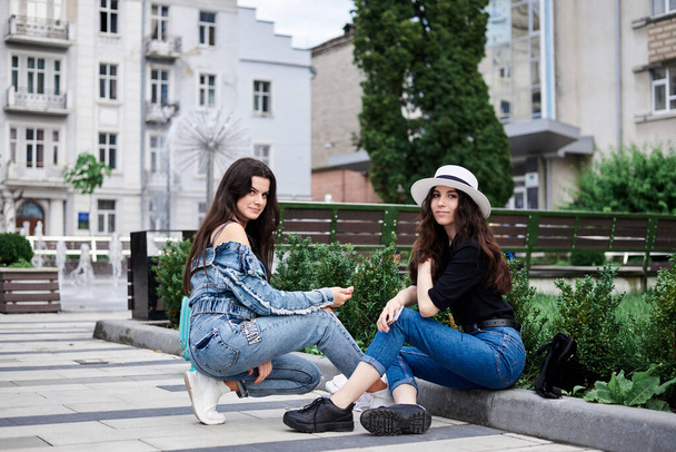 Two young brunette girls, wearing casual jeans clothes, with mint luggage, sitting on pavement border, checking phone in front of historical building in town. City sightseeing tour.Tourism lifestyle. - Foto, afbeelding