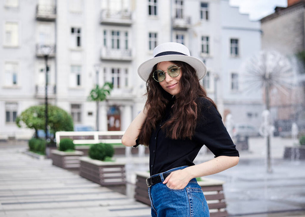 Young brunette woman, wearing black shirt, jeans and white hat with green sunglasses, posing in front of fountain in old city town in summer. Traveler in city. Female photo session. - Photo, Image