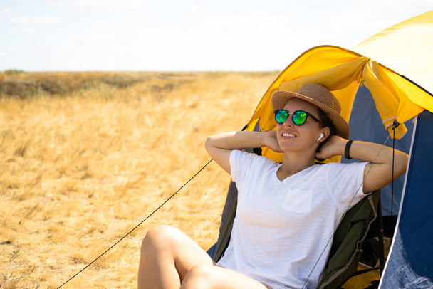 Ευτυχισμένη νεαρή γυναίκα τουρίστρια σε γυαλιά ηλίου, καλοκαιρινό καπέλο και ασύρματα ακουστικά χαλαρωτική μουσική ακρόασης, κάθεται μπροστά από σκηνή κάμπινγκ, να απολαύσετε την περιπέτεια κοντά στη λίμνη σε κίτρινο φόντο τομέα την ηλιόλουστη μέρα. - Φωτογραφία, εικόνα
