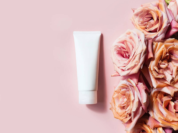 Κάτοψη της μακιγιαρισμένης φιάλης από λευκό πλαστικό σωλήνα και ροζ χρυσά τριαντάφυλλα σε ροζ φόντο σε σκόνη. Μπουκάλι για μαρκάρισμα και ετικέτα. Πολυτελή καλλυντικά. Αντιγράψτε χώρο, Flower flatlay - Φωτογραφία, εικόνα