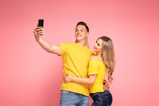 Excited young couple, two friends posing on pastel pink background studio, doing selfie shot on mobile phone. - Foto, Bild