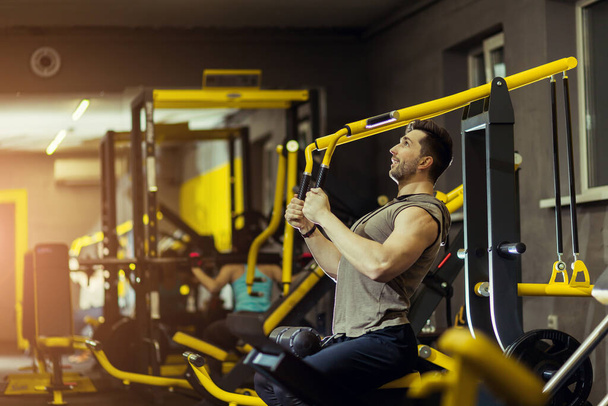 Мощный мускулистый человек, тренирующийся в тренажерном зале, делая упражнения на тяге на тренажере - Фото, изображение