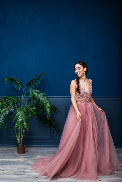młoda piękna kobieta w długiej różowej sukni wieczorowej, zmysłowa, uśmiechnięta, luksusowy styl przed niebieską ścianą i palmą - Zdjęcie, obraz