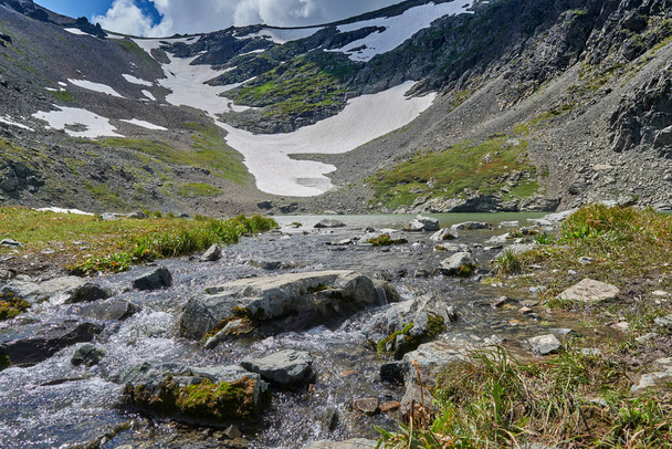 ορεινό ποτάμι που ρέει από μια λίμνη που σχηματίζεται από έναν παγετώνα ψηλά στα βουνά. Ωραία θέα στο βουνό και στις πλαγιές των βουνών με χιόνι - Φωτογραφία, εικόνα