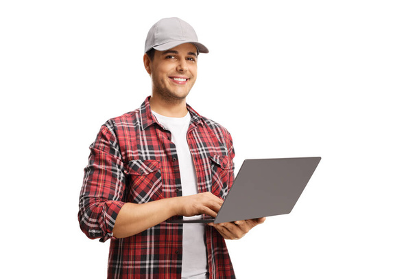 Young man with a cap holding a laptop computer and smiling at the camera isolated on white background - 写真・画像