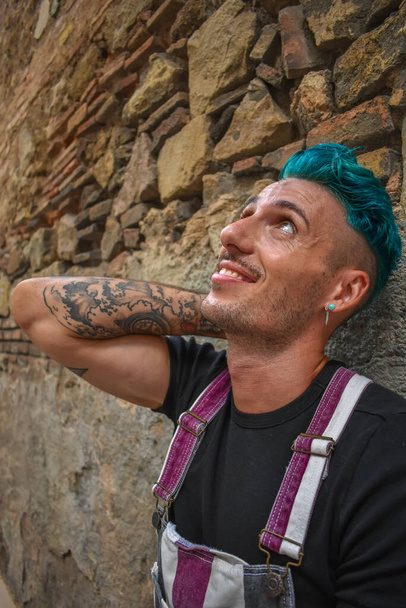 Young man wearing overalls, with blue hair, leaning against an old brick wall - Photo, Image