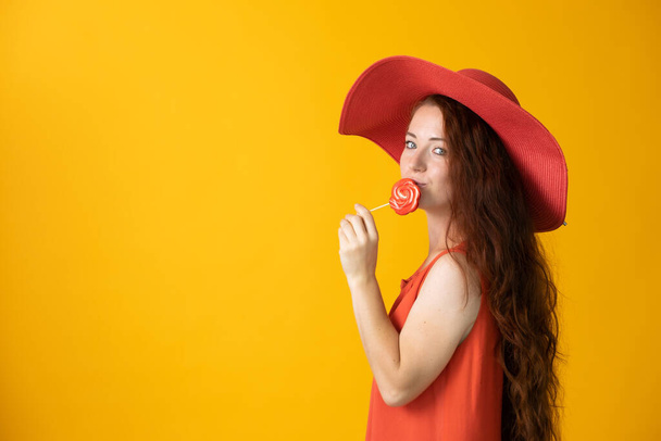 beautiful woman with long hair in a big red hat and lollipop on a yellow backgroun - Valokuva, kuva