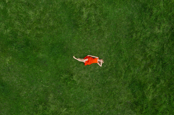 lonely blonde woman in red dress lying on green grass closing her face with hand shy excitement shame emotions. feminine figure. symbolic metaphor. Conceptual aerial artistic photo Complementary color - Photo, Image