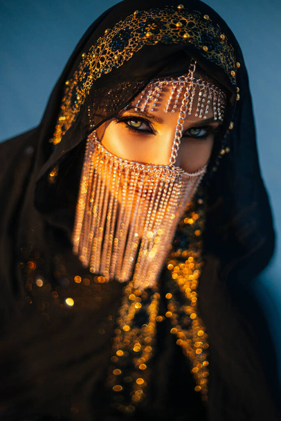 portrait of young beautiful oriental mysterious girl. The woman looks into the camera. The face is hidden by a golden veil. Luxurious evening make-up, smoky eyes. The head is covered with black scarf - Фото, зображення