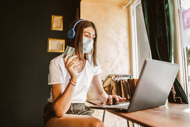 Casual woman in face mask, headphones using laptop and credit card for online shopping in cafe - 写真・画像