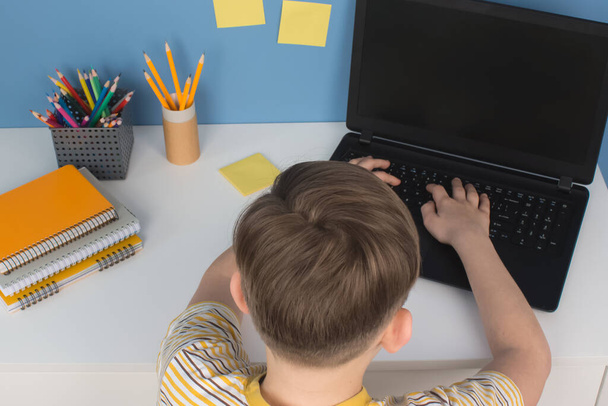 Boy using laptop computer for online studying during quarantine, sitting at the table agains blue wall. Homeschooling, online training classes or education technology concept - Photo, image
