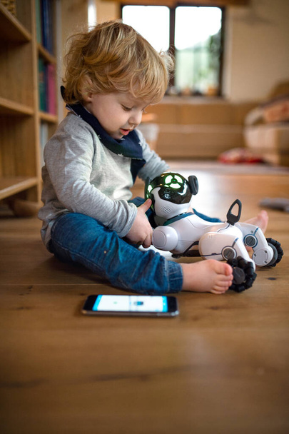 A cute toddler boy sitting indoors at home, playing with robotic dog. - Foto, imagen