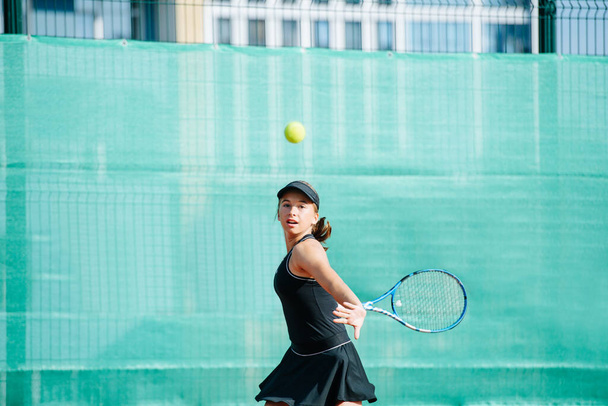 Teenage girl in a black sportive outfit playing tennis on a brand new court, returning ball fast. Over a banner covering fence and high storey apartment buildings - Photo, Image