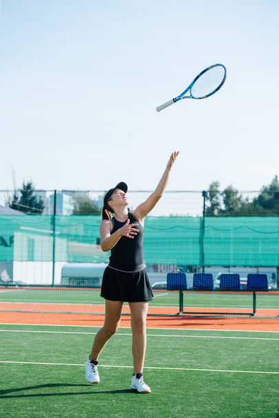Teenage girl in a black sportive outfit training on a new tennis court, playing with the racket, throwing it up in the air. Over a banner covering fence and high storey building. - 写真・画像
