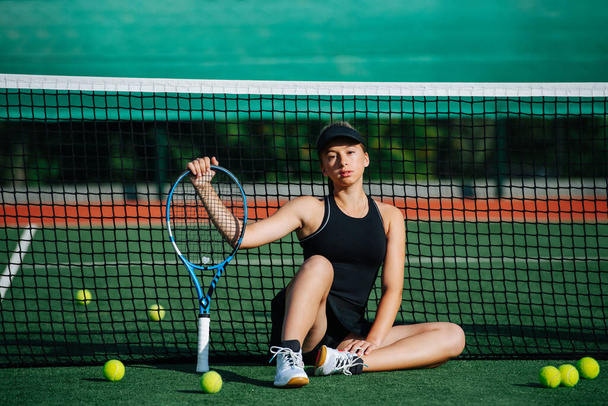 Teenage girl in a sportive outfit sitting in the middle of a brand new tennis court on a sunny day. She's wearing short tennis skirt with top and cap. Leaning on the net, looking at the camera. - Foto, imagen