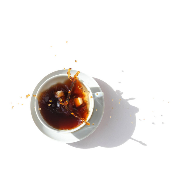 Fekete kávé fehér pohárban, felülnézetből. Cukorcsomókat dobnak a pohárba, látványos fröccsenések ömlenek ki a pohárból. Fehér háttér. Nemzetközi kávénap. - Fotó, kép