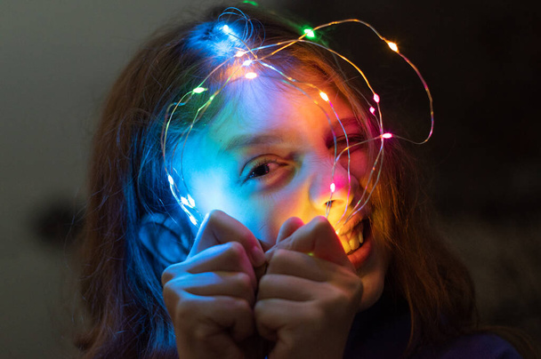 A little girl looks through a multi-colored garland. - Foto, afbeelding
