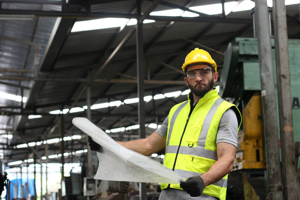 Προσωπογραφία του βιομηχανικού μηχανικού που φοράει κράνος και γυαλιά ασφαλείας που στέκονται και κρατούν το σχέδιο, που εργάζονται στο εργοστάσιο της βιομηχανίας παραγωγής - Φωτογραφία, εικόνα