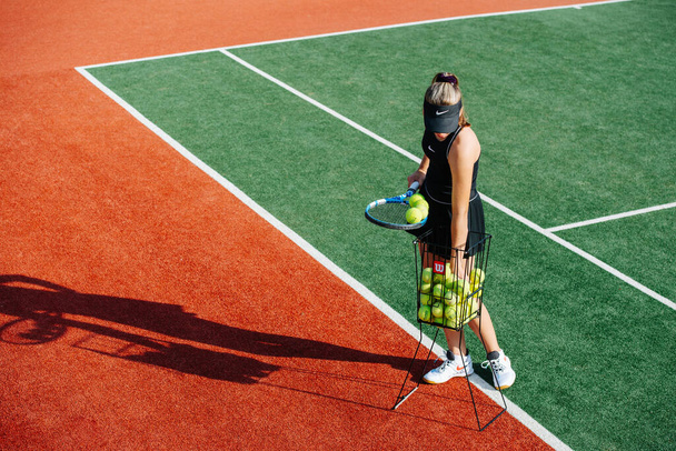 Teenage girl in a black sportive outfit standing on a tennis court on a sunny day. She is taking balls from a basket for her practice - Фото, зображення