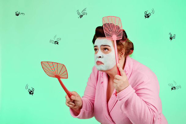 nadváha legrační žena na sobě růžový župan, vlasy natáčky a zelená maska krásy honí mouchy s plácačkou na zeleném izolovaném pozadí - Fotografie, Obrázek