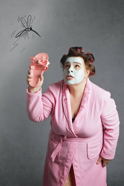 nadváha legrační žena na sobě růžový župan, kadeře a zelená maska krásy honí komára s pantoflem na šedém izolovaném pozadí - Fotografie, Obrázek