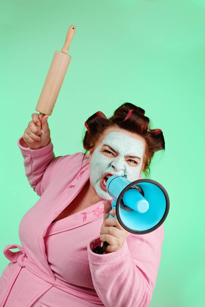 overweight funny woman wearing pink bathrobe, hair curlers and green beauty mask shouting into megaphone on isolated green background - Photo, Image