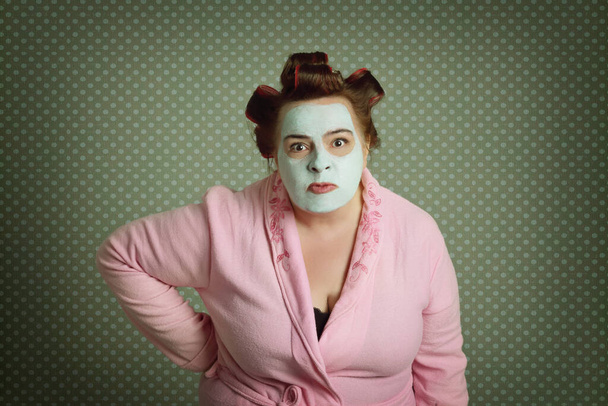 retrato de irritado excesso de peso mulher engraçada vestindo roupão de banho rosa, encrespadores de cabelo e máscara de beleza verde isolado em um fundo cinza  - Foto, Imagem