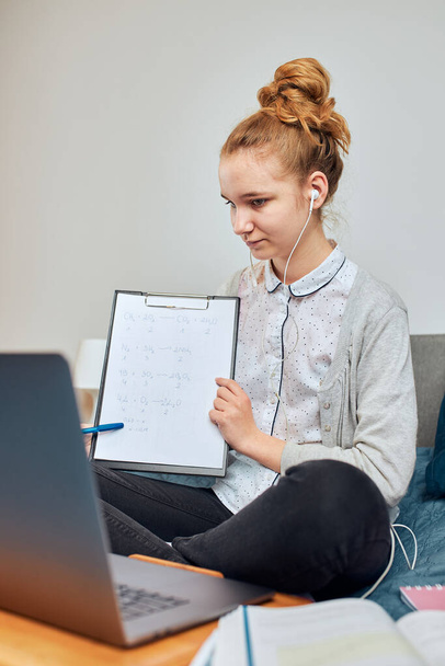 Young woman student having classes, learning online, watching lesson remotely, listening to professor, talking with classmates on video call from home during quarantine. Young girl using laptop, headphones, books, manuals sitting on bed - Фото, изображение