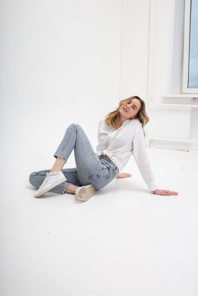 donna caucasica posa in camicia e jeans blu, seduta sul pavimento bianco dello studio - Foto, immagini
