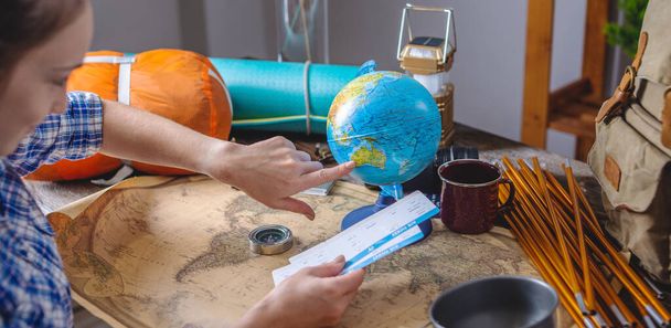 A woman is choosing a place for the next trip on the globe at a wooden table with camping equipment . Concept of travel and discovery - Photo, Image
