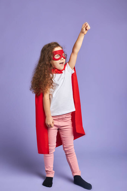 Cheerful little girl wearing red mask and cloak isolated purple background - 写真・画像