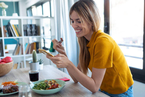 Prise de vue d'une femme souriante mangeant des aliments sains en utilisant son téléphone portable à la maison. - Photo, image