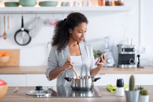Shot of confident young african american woman cooking healthy food while using her mobile phone in the kitchen at home. - Valokuva, kuva
