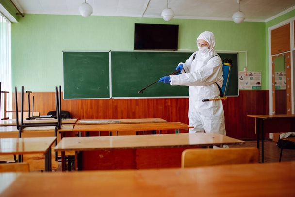 Mies suihkuttaa desinfiointinestettä luokkahuoneen pöydille ennen koulukautta. Saniteettityöntekijä puhdistaa auditorion suojapuvussa. Opiskelijat ja oppilaat terveydenhuollon käsite - Valokuva, kuva