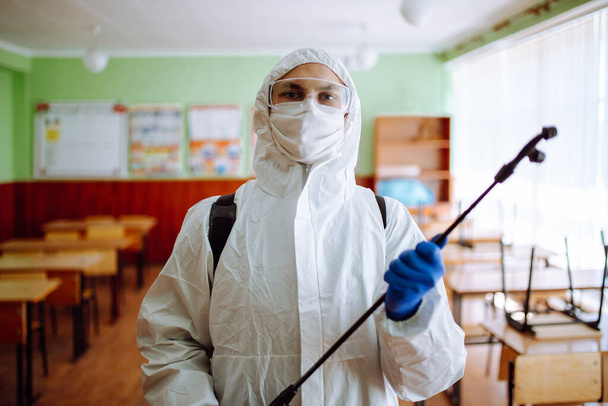 Πορτρέτο ενός άνδρα με προστατευτική αντιβακτηριακή στολή που καθαρίζει την τάξη με ένα σπρέι με απολυμαντικό υγρό. Επαγγελματίας υγειονομικός απολυμαίνει το αμφιθέατρο με ειδικό εξοπλισμό - Φωτογραφία, εικόνα