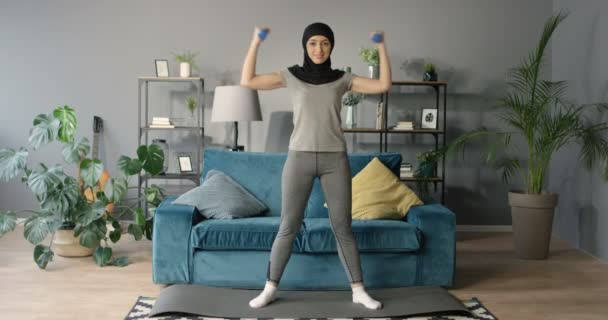 黒のスカーフでかなりスポーティな若いアラビア人女性は自宅でダンベルと強い腕のための運動をしています。美しいスリムムスリム女性の運動やリビングルームでの上作業. - 映像、動画