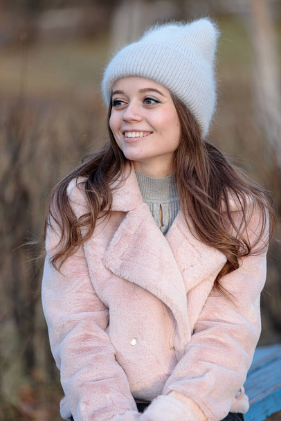 Een jong mooi meisje met een grijze pluizige hoed en een roze jas kijkt weg met een glimlach. - Foto, afbeelding