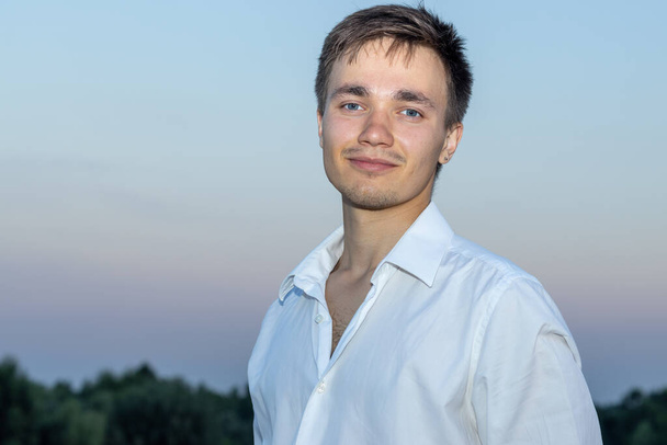 Portrait of a young man in a white shirt against the backdrop of the sunset sky. - Фото, изображение