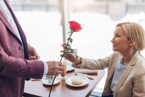 elegante man geeft een mooie roos aan glimlachende mooie blonde vrouw in cafe - Foto, afbeelding
