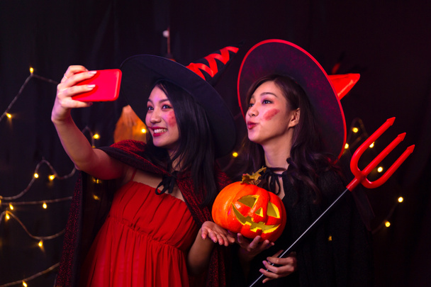 Dos mujeres tomando fotos con teléfono móvil en la fiesta de Halloween. Selfie con teléfono móvil en la fiesta. Una mujer vestida de bruja y un demonio sosteniendo una calabaza. - Foto, imagen