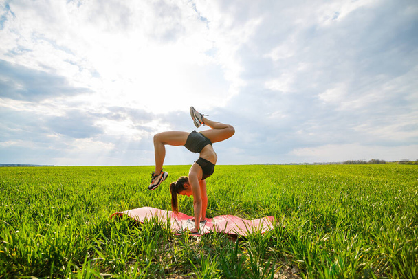 Beautiful young woman in a black top and shorts performs a handstand. A model stands on her hands, doing gymnastic splits against the blue sky. Healthy lifestyle concept - Photo, Image