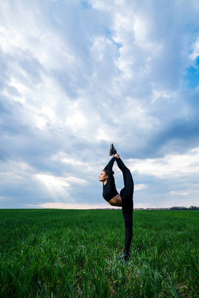 krásná dívka gymnastka na zelené trávě dělat jógu. Krásná mladá žena na zeleném trávníku provádí akrobatické prvky. ohebné gymnastka v černé dělá handstand v split - Fotografie, Obrázek