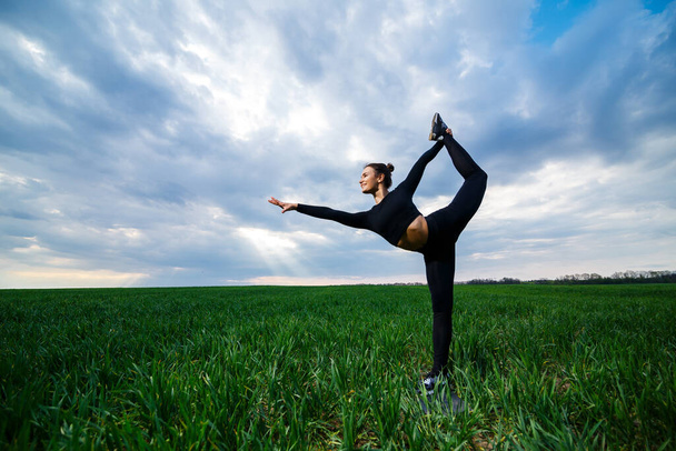 Yeşil çimlerin üzerinde güzel bir jimnastikçi kız yoga yapıyor. Yeşil çimenlikteki güzel genç bir kadın akrobatik öğeler sergiliyor. Siyahlı esnek jimnastikçi ikiye bölünmüş amuda kalkar - Fotoğraf, Görsel