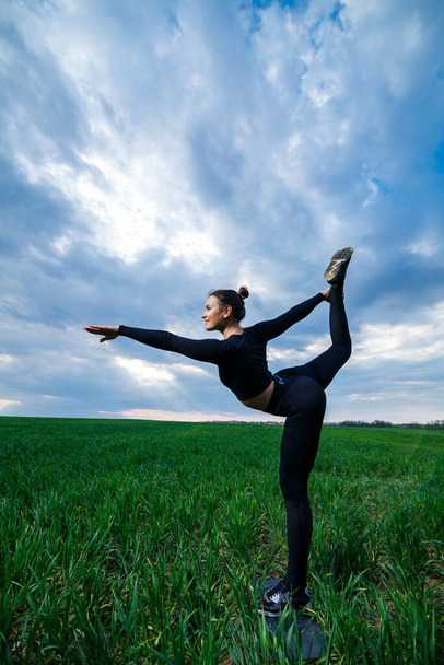 mooie meisje gymnaste op het groene gras doen yoga. Een mooie jonge vrouw op een groen gazon voert acrobatische elementen uit. flexibele turner in het zwart doet een handstand in split - Foto, afbeelding