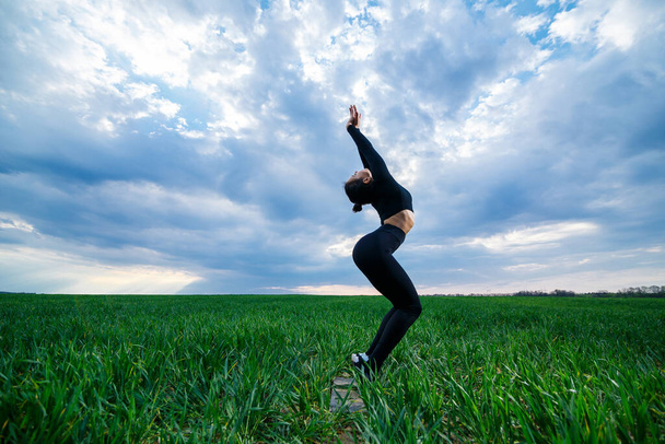 beautiful girl model on green grass do yoga. A beautiful brunette on a green lawn performs acrobatic elements. flexible gymnast in black is doing exercises - Photo, Image