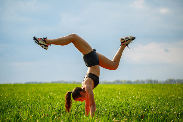 beautiful girl model on green grass do yoga. A beautiful young woman on a green lawn performs acrobatic elements. flexible gymnast in black does a handstand in split - Φωτογραφία, εικόνα