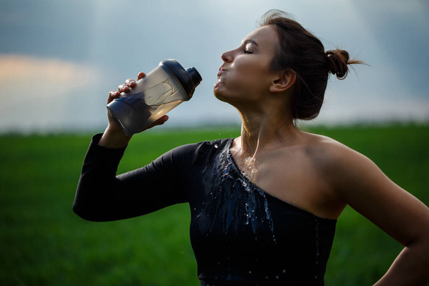 Όμορφη μελαχρινή κοπέλα, με μακριά μαλλιά στον άνεμο, πίνοντας από ένα μπουκάλι νερό. Εξωτερικές πόρτες, μπλε φόντο του ουρανού. - Φωτογραφία, εικόνα