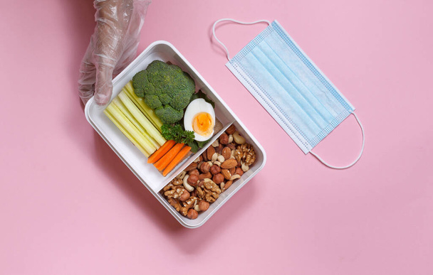 Mãos em luvas protetoras com um almoço útil em uma caixa e uma máscara protetora. Visão horizontal superior sobre um fundo rosa - Foto, Imagem